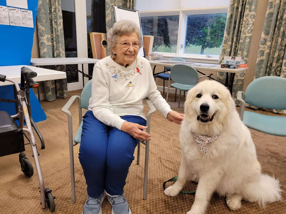 An elderly woman sitting on a chair indoors, smiling and petting a large white fluffy dog wearing a patterned bandana. The room has carpeted flooring, several chairs, a walker, a table with board games and puzzles, and windows with curtains showing greenery outside.