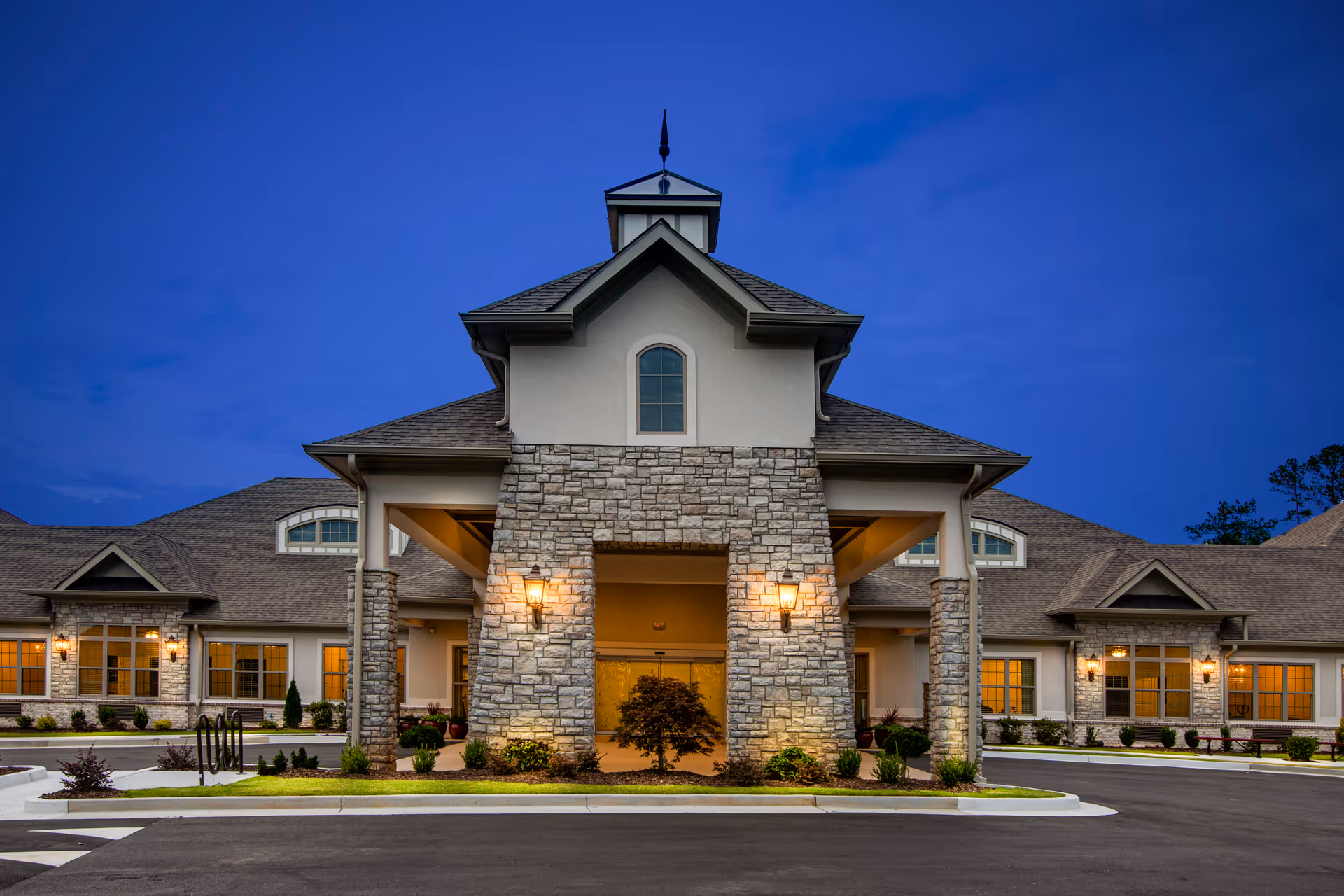 Stone-faced main entrance of a senior living building lit at dusk with lanterns and a circular driveway.
