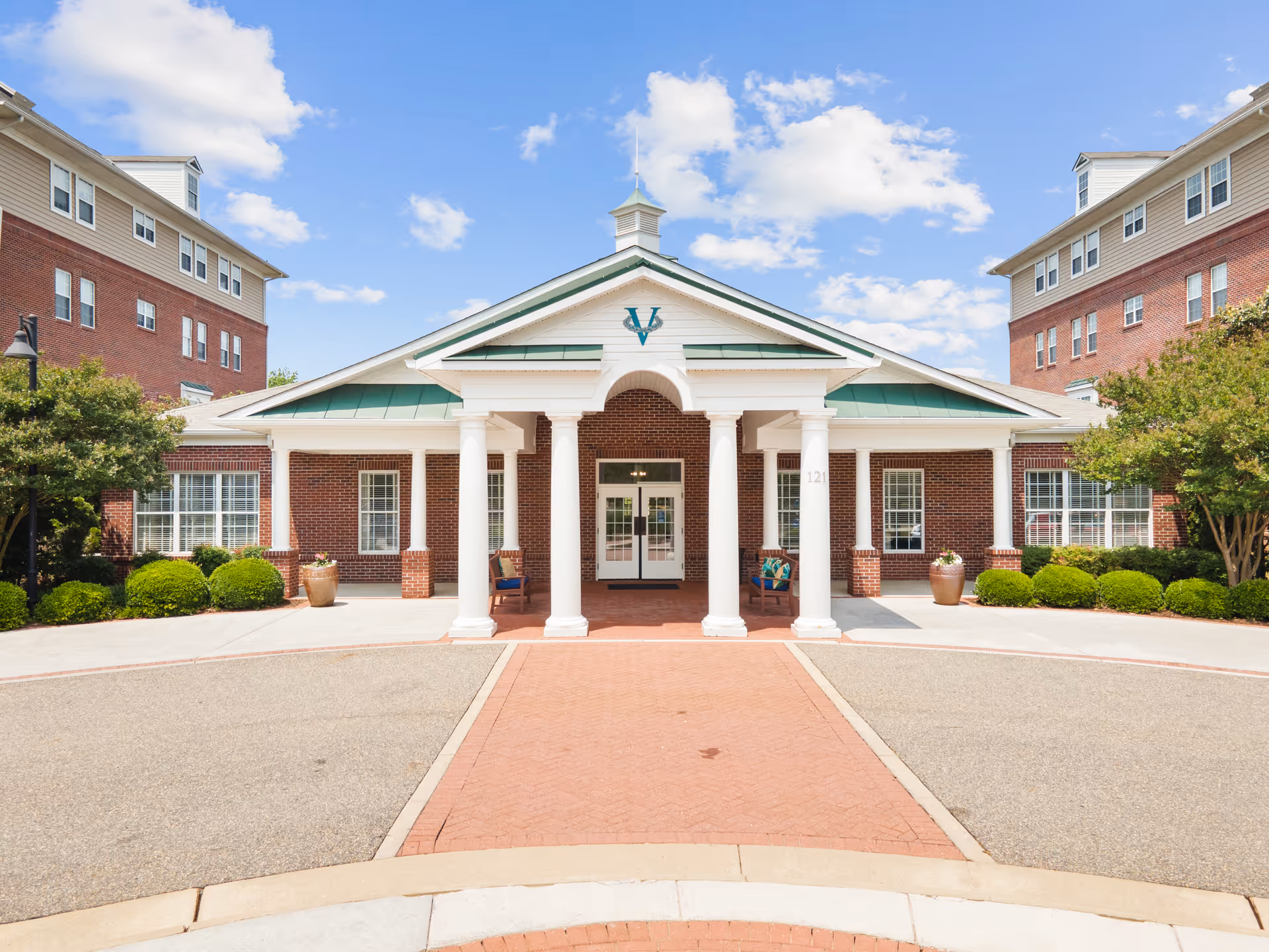 Front exterior view of Verena at the Reserve facility showing a brick building with white columns supporting a green roofed entrance. There are shrubs and trees on either side of the entrance, and a clear blue sky with some clouds above.