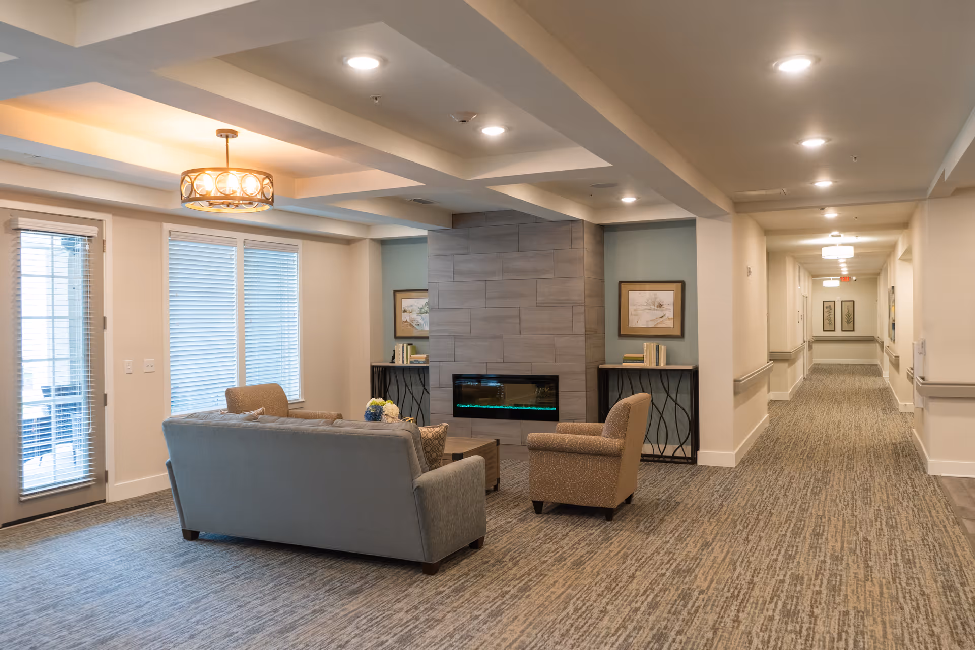 A cozy common area in a senior living facility featuring a gray sofa and an armchair arranged around a modern electric fireplace set in a tiled wall. The room has soft lighting from ceiling fixtures and a decorative hanging light. There are two windows with blinds and a door leading outside. The space includes side tables with books and framed artwork on the walls. A long hallway extends from the room, lined with handrails and additional framed pictures.
