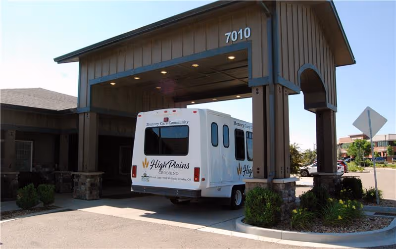 A High Plains Crossing shuttle van is parked under the covered entrance of a senior living building numbered 7010.
