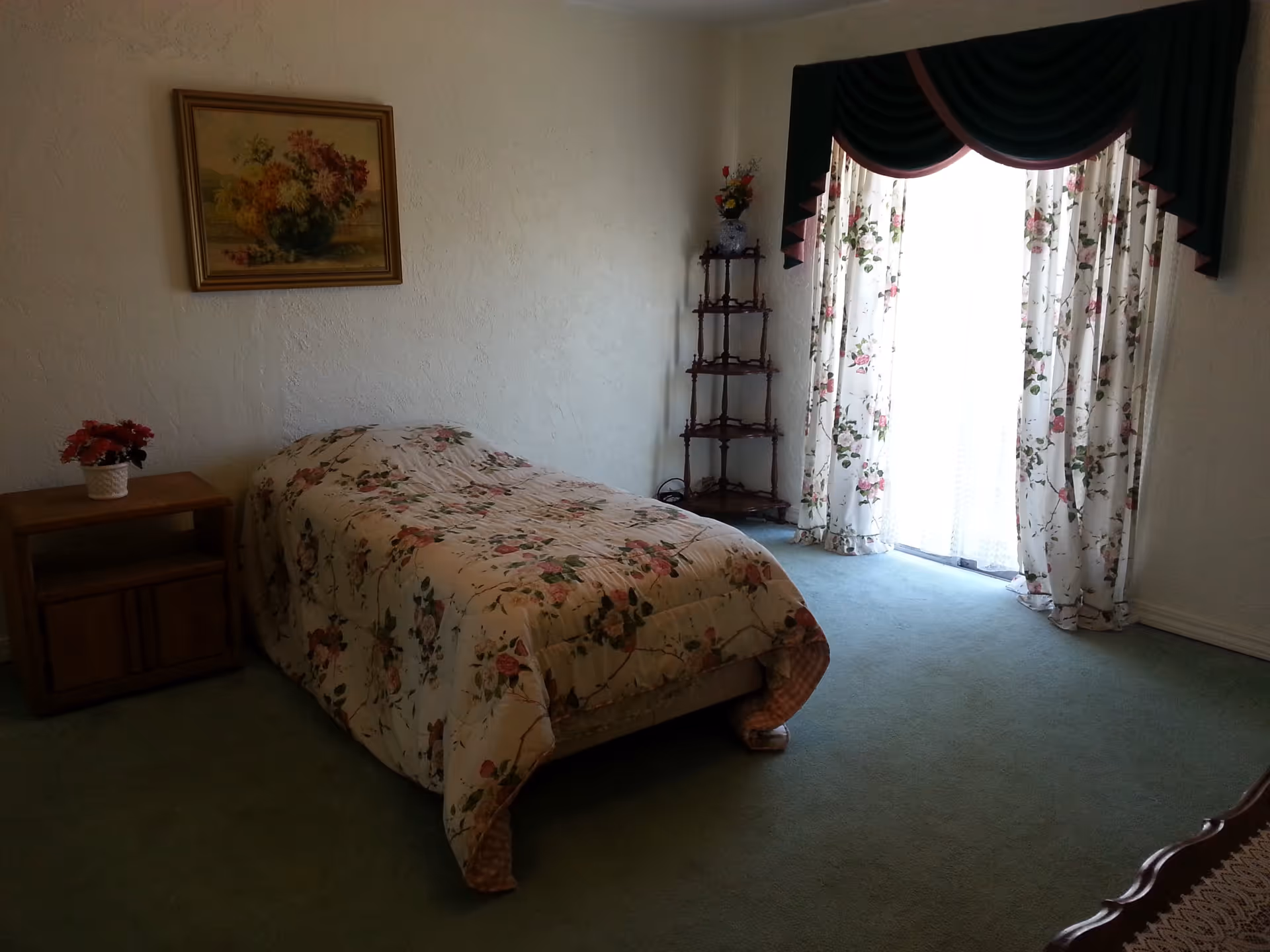 A bedroom with a single bed covered in a floral bedspread, a wooden nightstand with a small flower pot, a corner shelf with a vase of flowers, floral curtains with a dark green valance, and a framed painting of flowers on the wall.