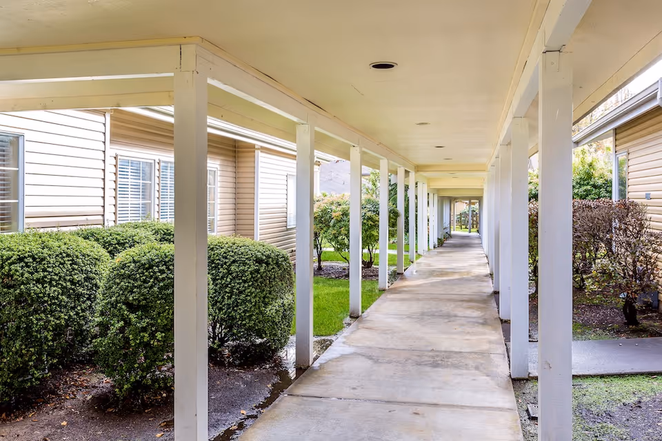 Covered outdoor walkway between beige cottages lined with white columns, windows, and trimmed shrubs.