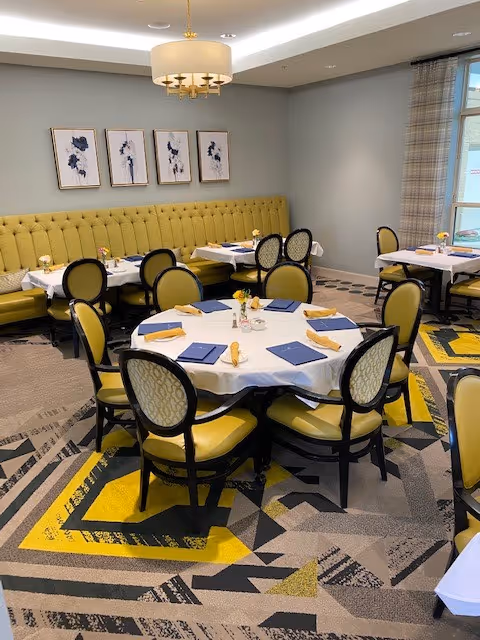 Dining room with round and rectangular tables covered with white tablecloths, set with blue napkins and yellow rolled napkins. Yellow cushioned chairs surround the tables, and a long yellow upholstered bench lines the wall. Three framed abstract floral artworks hang above the bench, and a modern chandelier is mounted on the ceiling. A window with plaid curtains is visible on the right side.