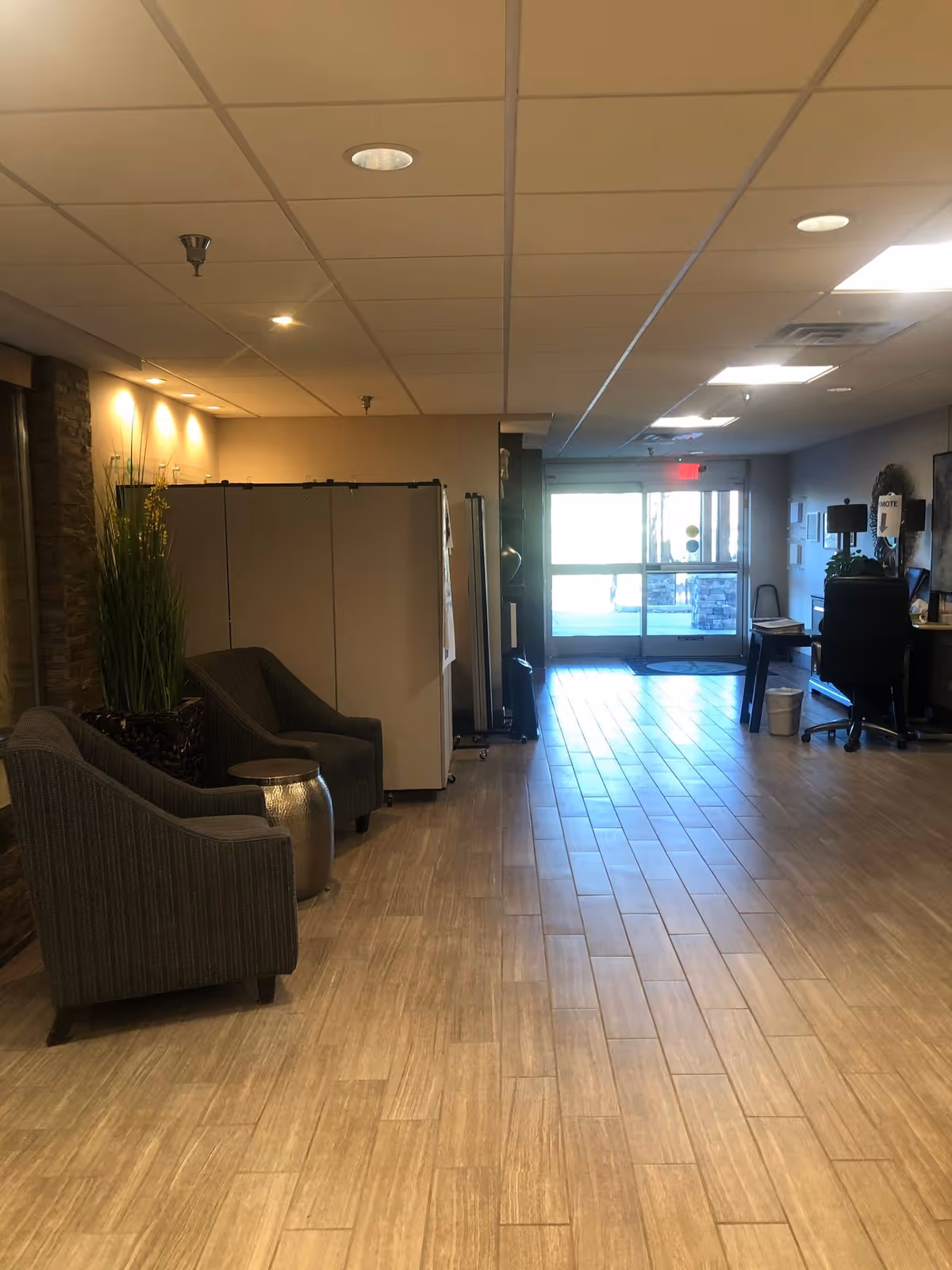 Interior view of a facility hallway with two gray armchairs and a small metallic side table on the left, a tall plant in a pot, and a reception desk with a chair on the right near the entrance door with glass panels letting in natural light.