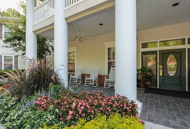 Front porch area of a building with large white columns, a green door with an oval window, three white rocking chairs, ceiling fan, and a garden with various colorful flowers and plants in the foreground.