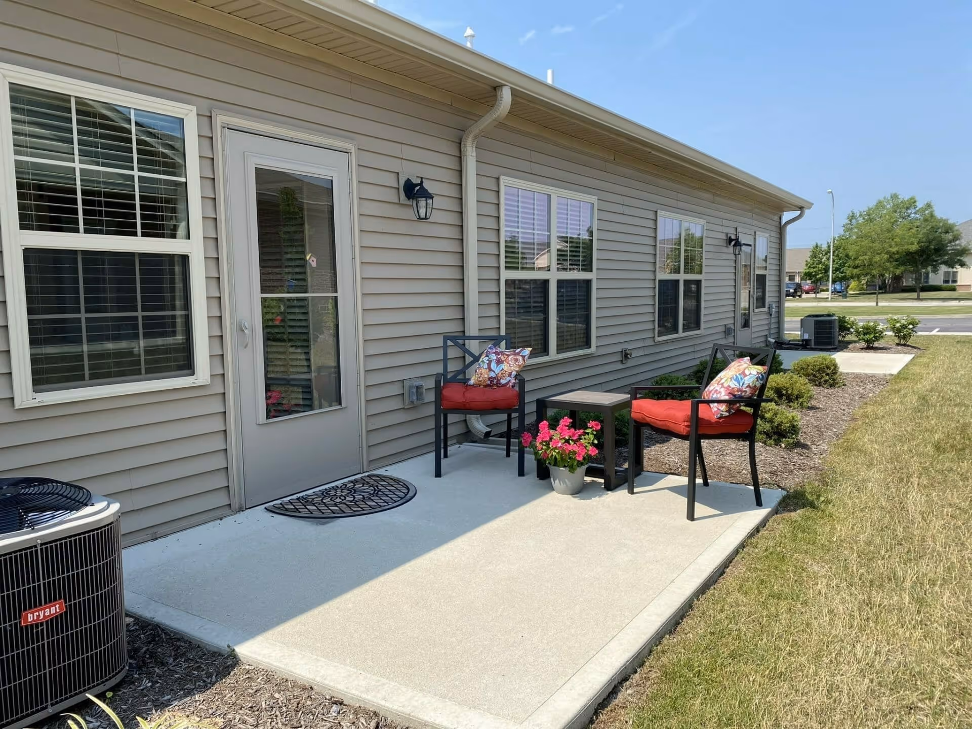 Outdoor patio area of a senior living facility with two black metal chairs with red cushions and colorful pillows, a small black table, and a potted pink flower. The patio is adjacent to a beige siding building with several windows and a glass door. There is an air conditioning unit on the left side and a grassy lawn on the right.