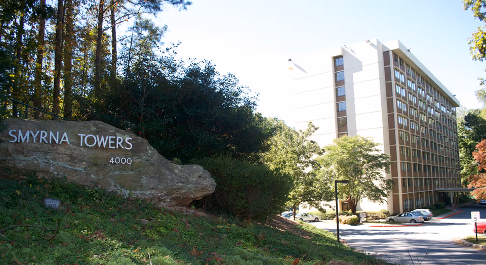 Exterior view of Smyrna Towers Senior Apartments, showing a multi-story building surrounded by trees and a parking area. A large rock with the name 'Smyrna Towers' and the number '4000' is visible in the foreground on a grassy slope.