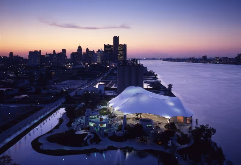 Aerial view of a city skyline at dusk with a large river on the right side. In the foreground, there is a uniquely shaped white tent-like structure illuminated with lights, surrounded by water features and greenery. The sky is a gradient of purple and orange as the sun sets.