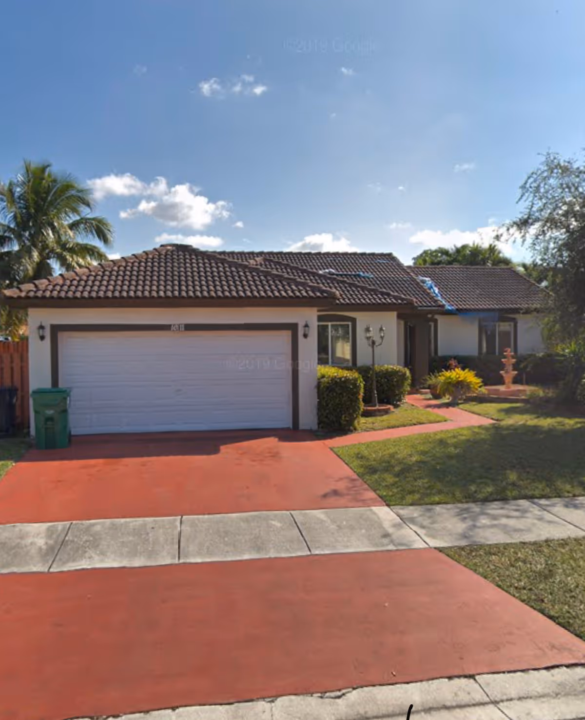Single-story house with a red driveway, a two-car garage with a white door, and a tiled roof. There are bushes and a small garden with a fountain on the right side of the house. A palm tree is visible on the left side, and the sky is clear with a few clouds.