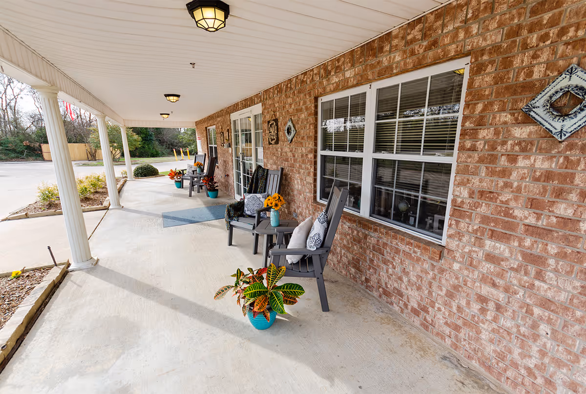 Covered outdoor porch area with brick walls, white ceiling, and columns. Several black chairs with cushions and small tables with potted plants and flowers are arranged along the wall near windows and a glass door. The porch overlooks a parking area and some greenery.