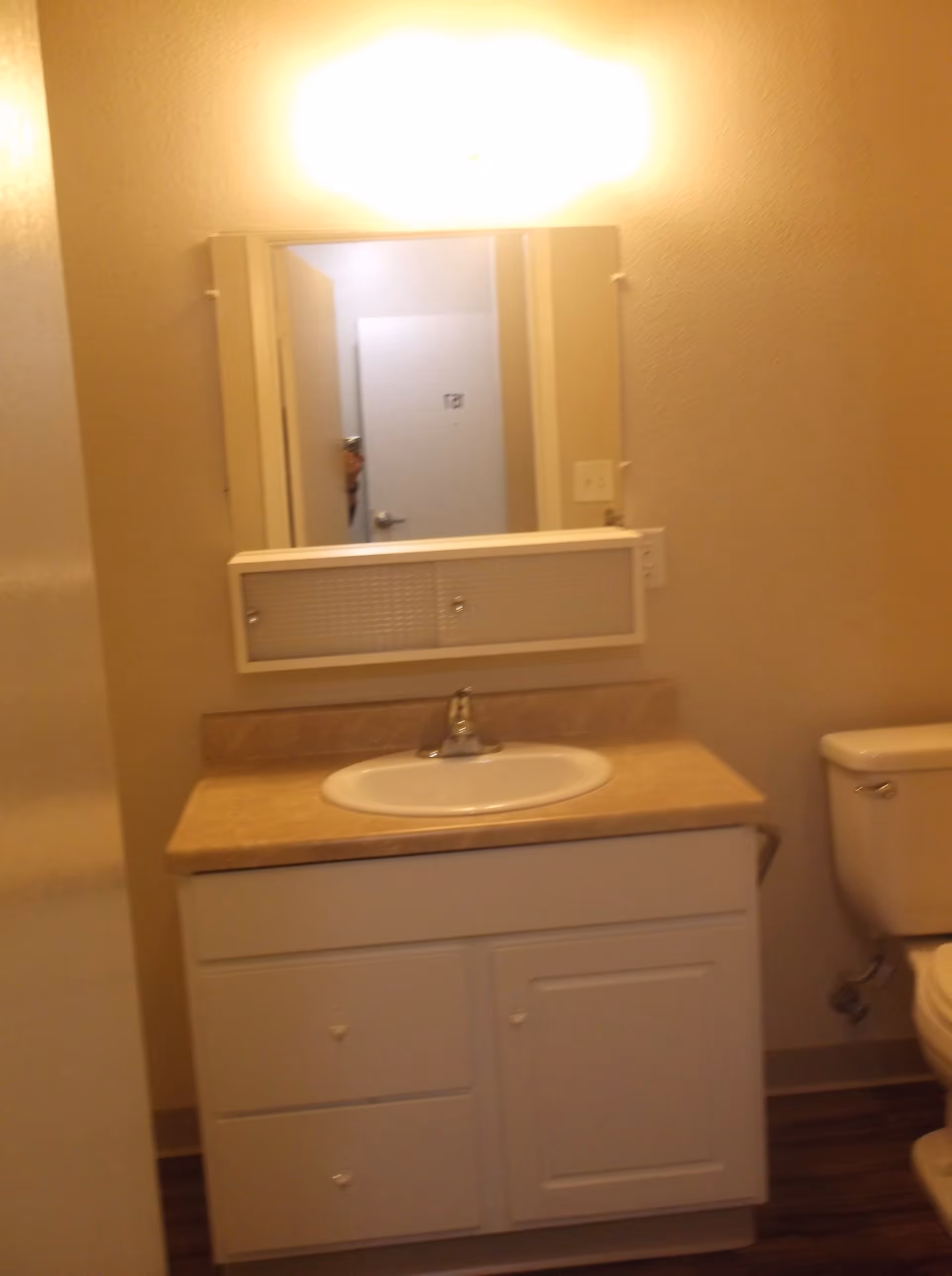 A small bathroom vanity with a beige countertop, white sink, and white cabinet below. Above the sink is a rectangular mirror with a light fixture above it. To the right of the vanity is a white toilet. The walls are painted a light beige color and the floor has a wood-like finish.