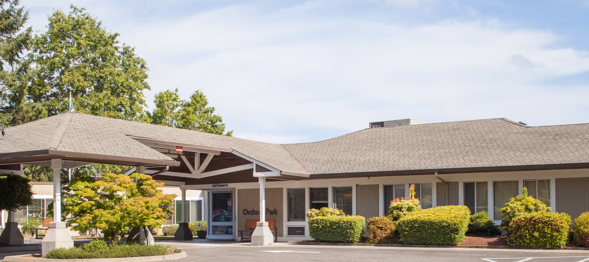 Exterior view of Orchard Park Health and Rehabilitation Center showing the main entrance with a covered drop-off area, surrounded by neatly trimmed bushes and trees under a partly cloudy sky.