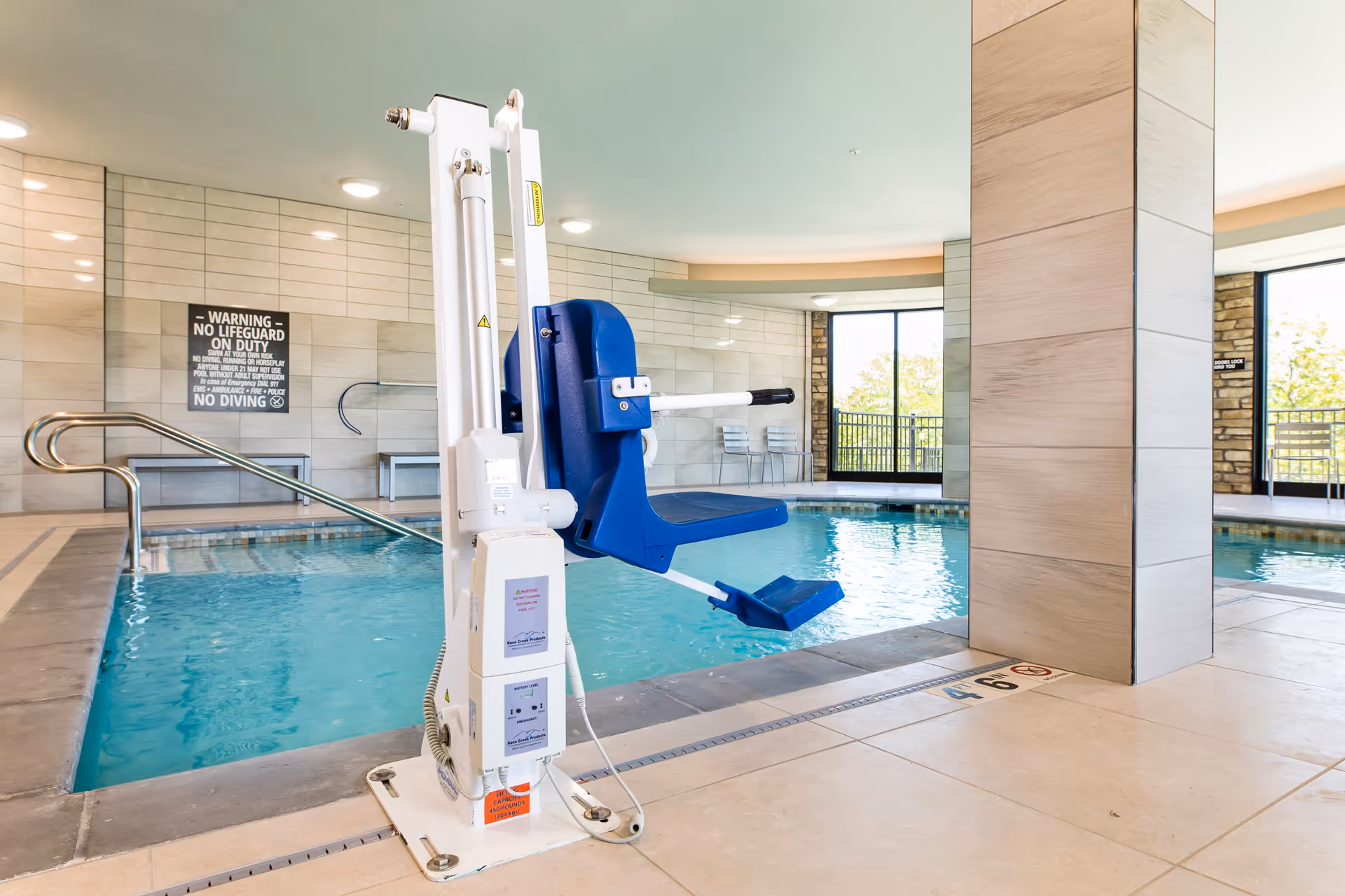 Indoor swimming pool area with a blue pool lift chair for accessibility. The pool is surrounded by tiled flooring and walls, with large windows letting in natural light. There is a warning sign on the wall stating no lifeguard on duty and no diving.