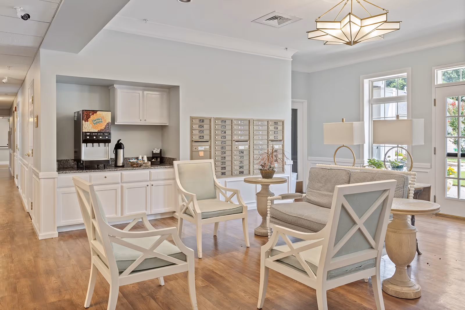 A bright and airy common area in an assisted living facility featuring a seating arrangement with four white wooden chairs with light cushions and a small sofa around two round wooden tables. Behind the seating area is a countertop with a beverage dispenser, coffee pot, and cups. On the wall above the countertop are multiple mailboxes. Large windows and a glass door allow natural light to fill the room, and a modern chandelier hangs from the ceiling.