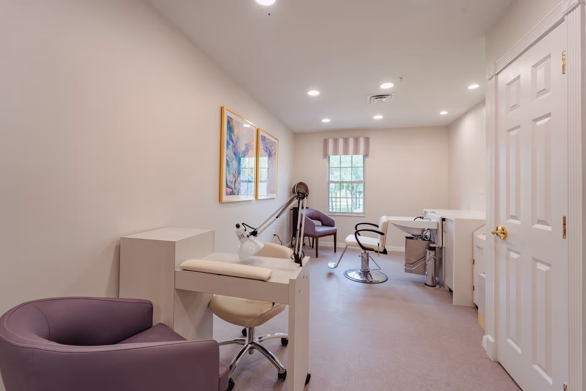 A well-lit room with a manicure table and lamp, a purple armchair, a salon chair in front of a sink, and a window with a striped valance. Two colorful framed artworks hang on the wall.