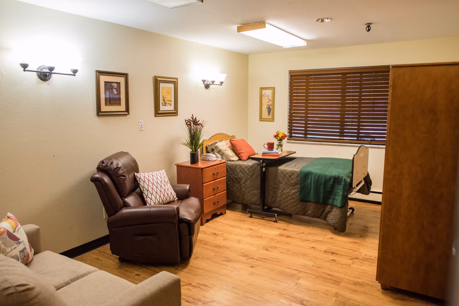 A cozy senior living bedroom with a single bed covered in a green quilt and pillows, a wooden nightstand with a plant and a telephone, a brown leather recliner chair with a patterned pillow, a beige sofa with a decorative pillow, wooden blinds on the window, framed artwork on the walls, and wooden flooring.
