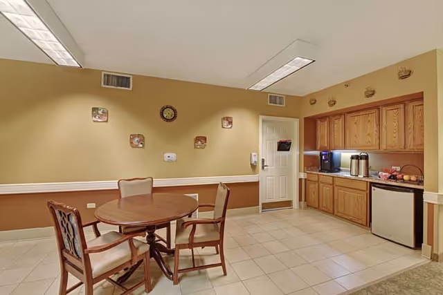 Small communal dining area with a round wooden table and chairs and a kitchenette with wood cabinets, coffee station and mini fridge against a beige wall.