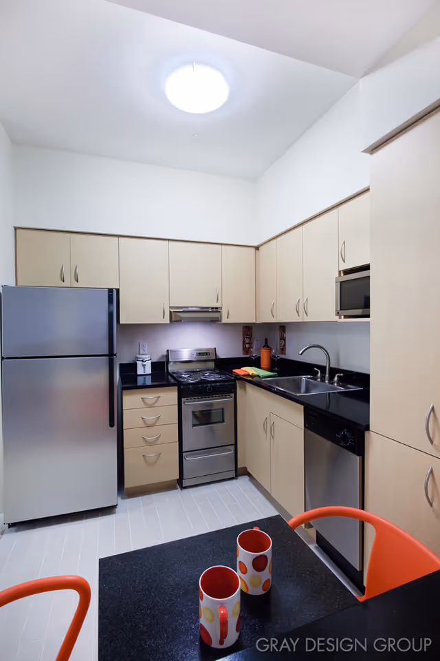 Modern kitchen with light wood cabinets, stainless steel refrigerator, stove, microwave, and dishwasher. Black countertops and a double sink are visible. In the foreground, there is a black table with two colorful mugs and orange chairs.
