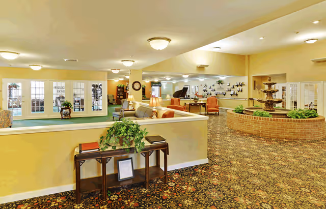 Spacious senior living facility common area with patterned carpet, beige walls, and ceiling lights. The room features a brick circular fountain with greenery, several seating areas with sofas and chairs, a piano, and decorative plants. French doors and large windows allow natural light into the space.