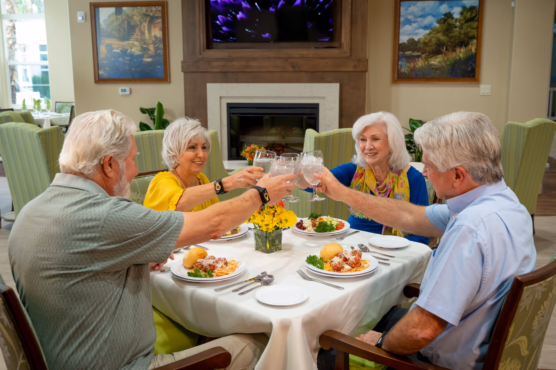 Four older adults seated at a dining table in a memory care facility, toasting with glasses over plated meals.