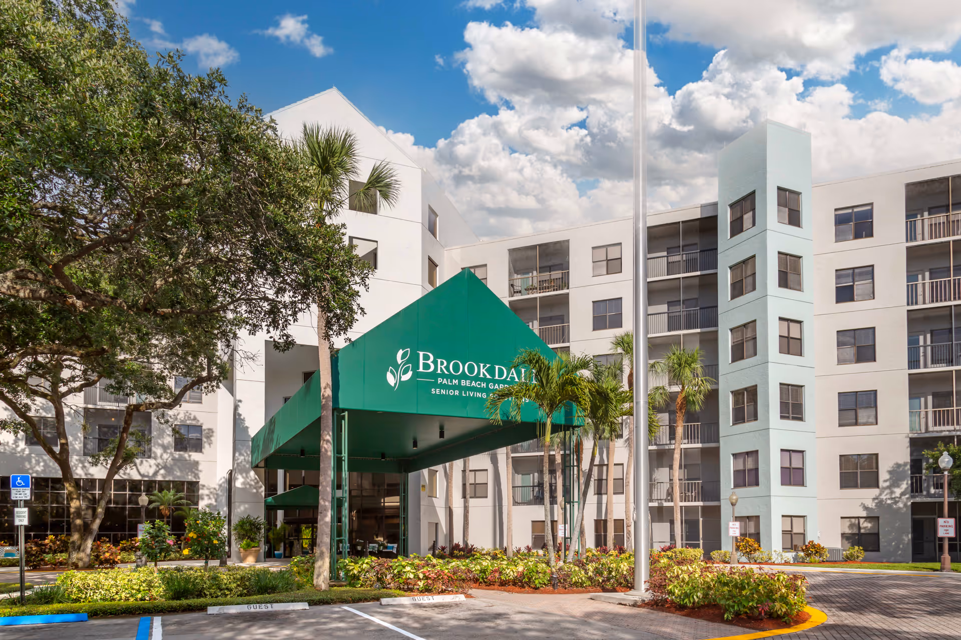Exterior view of Brookdale Palm Beach Gardens senior living facility showing a multi-story white building with balconies, a green entrance canopy with the facility name, palm trees, and landscaped greenery under a partly cloudy blue sky.