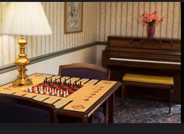 A cozy room with a table featuring a chess and backgammon board, two chairs, a brass lamp with a white lampshade, a piano with a yellow cushioned bench, and a vase with pink flowers on top. The walls have striped wallpaper and a framed picture.