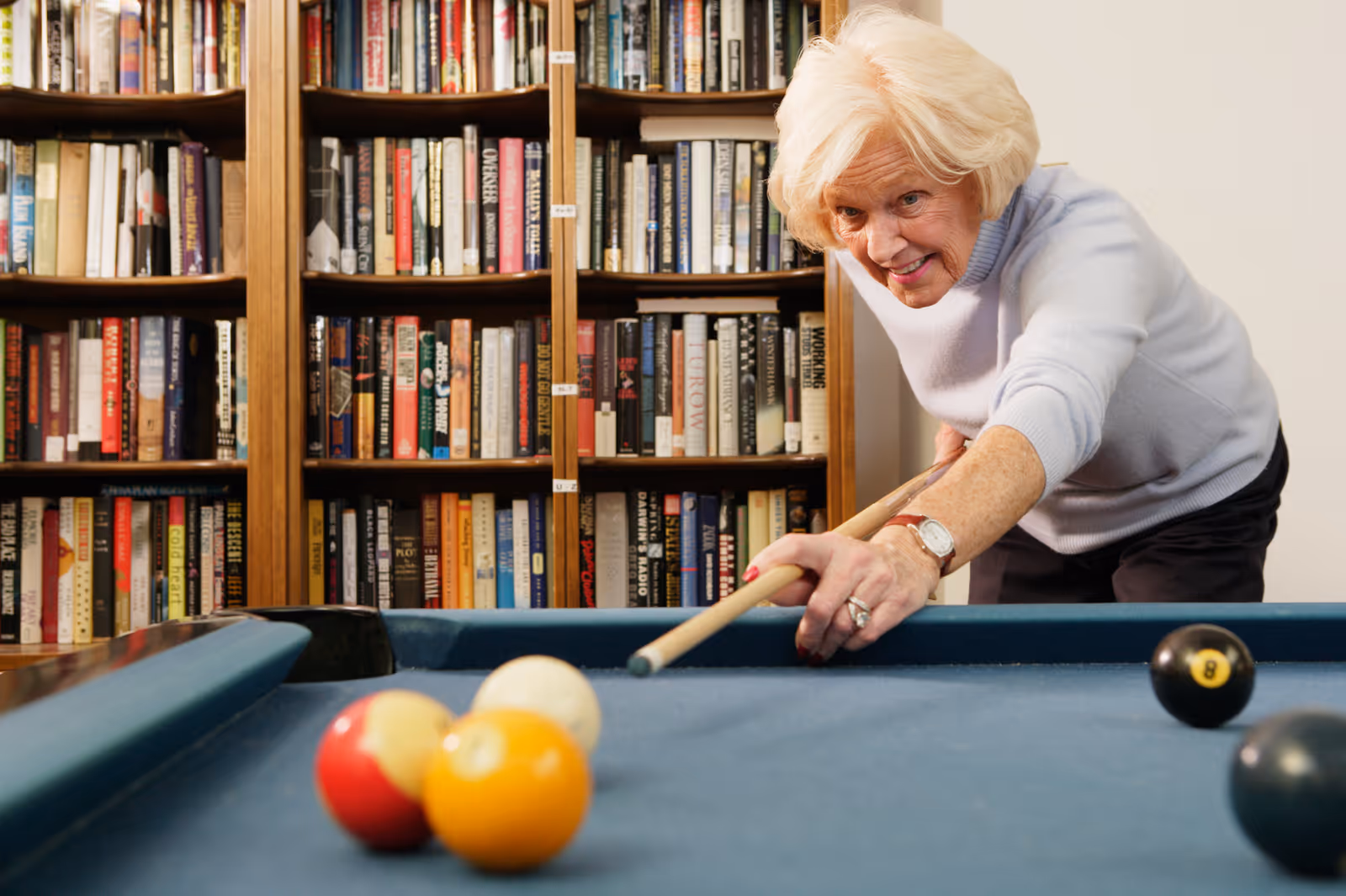 An elderly woman with white hair is playing pool, aiming her cue stick at the billiard balls on a blue pool table. Behind her is a large bookshelf filled with many books.