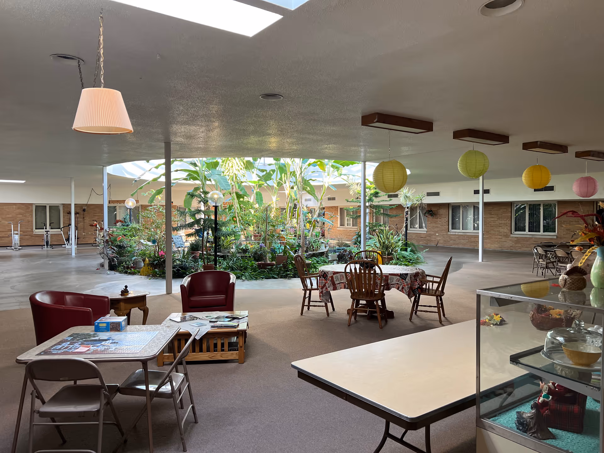 Open indoor common area with tables, chairs, and a central planted atrium beneath skylights.