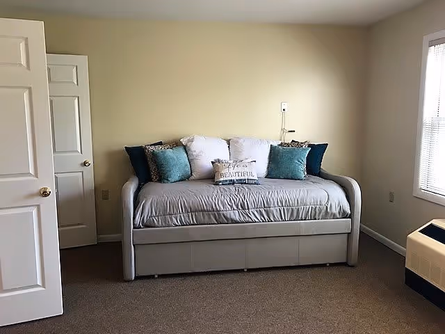 A simple room with a beige daybed against a light yellow wall. The daybed has a gray cover and several decorative pillows, including one with the words 'Life is Beautiful.' There is a window with blinds on the right side and an open door on the left side. The floor is carpeted in a neutral color.