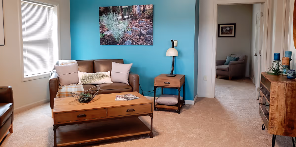 A cozy living room with a brown leather sofa adorned with pillows, a wooden coffee table with a decorative bowl and magazines, a side table with a lamp, and a bright turquoise accent wall featuring a nature-themed picture. There is a window with blinds on the left and a hallway leading to another room with an armchair and framed picture.