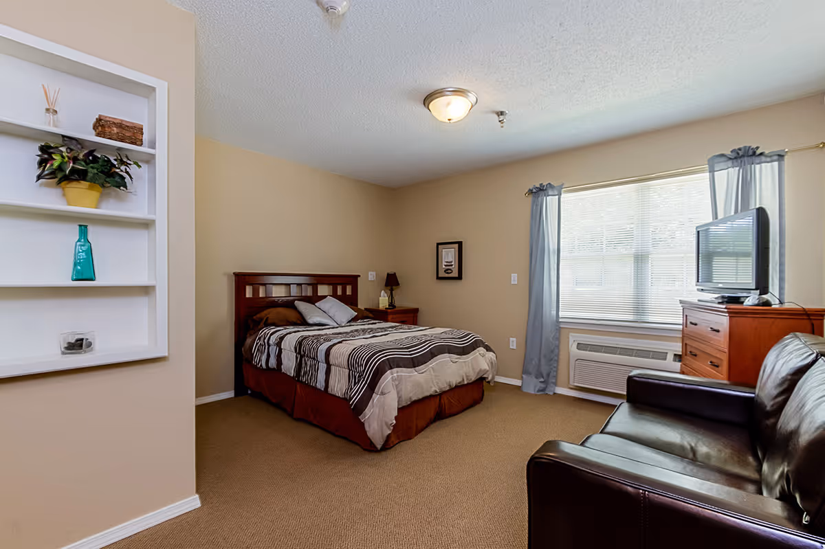A senior living facility bedroom with a bed covered in a striped brown and beige comforter, a wooden headboard, a nightstand with a lamp, a window with blue curtains, a wooden dresser with a TV on top, a brown leather couch, and a built-in wall shelf with decorative items.