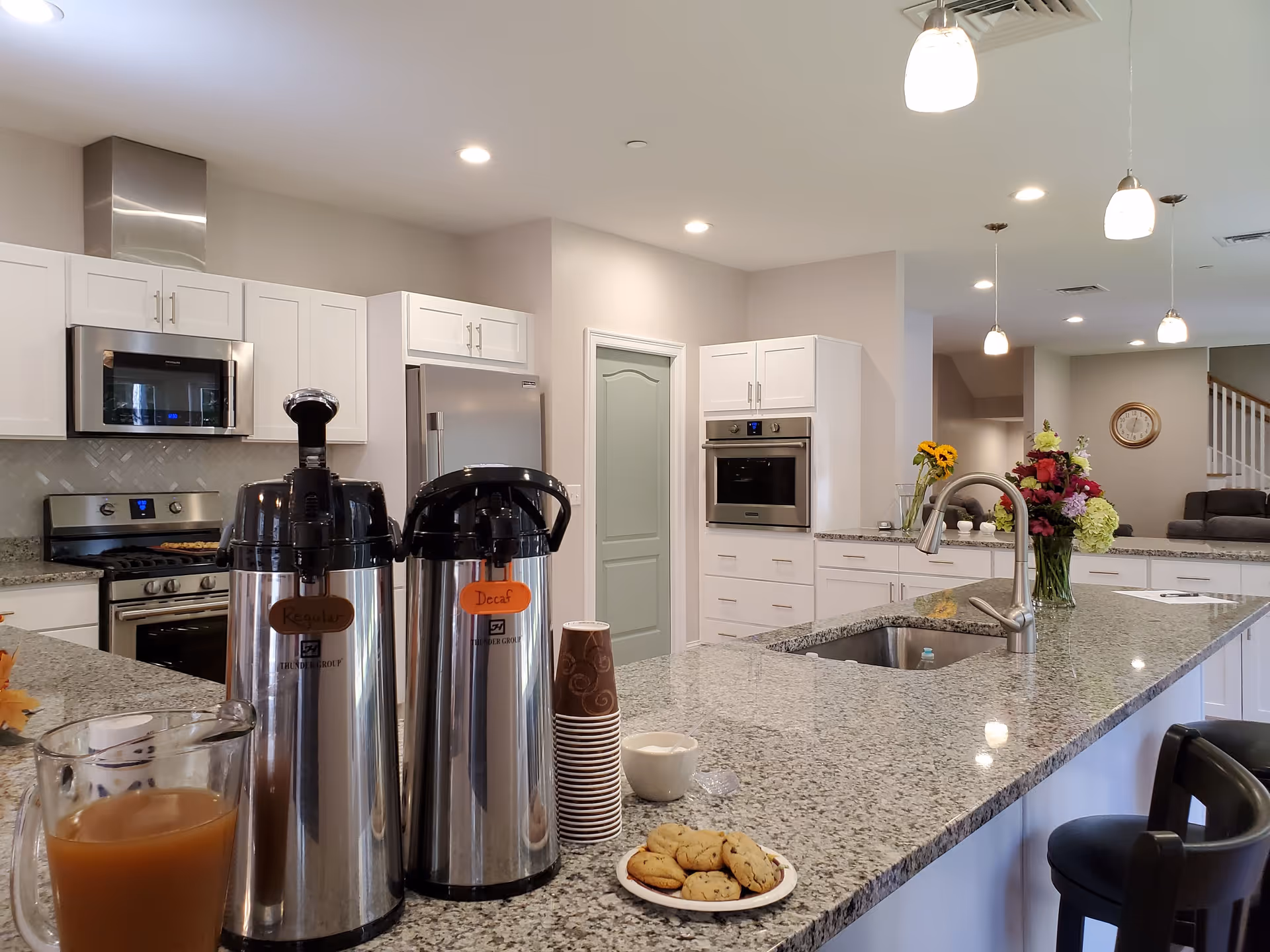 Spacious modern kitchen with a long granite island holding coffee dispensers, a pitcher, cookies, and a vase of flowers.