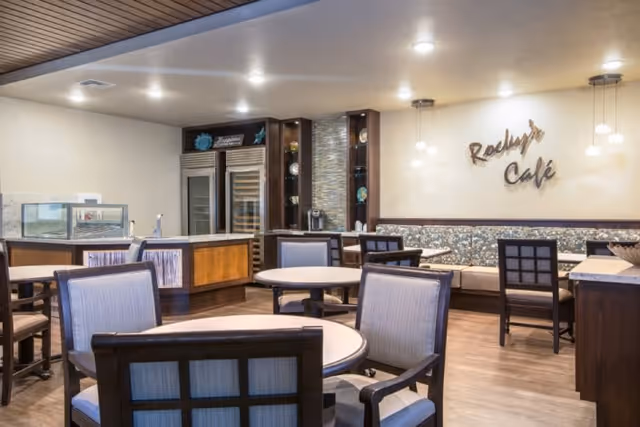 Interior view of a cozy café area named 'Rocky's Café' with round tables and cushioned chairs arranged neatly. The space features warm lighting, a wooden floor, and a counter with refrigerated display units and shelves in the background.
