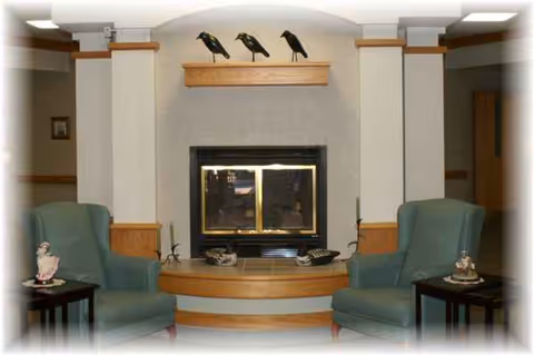 A cozy senior living common area with a centered fireplace, mantel birds, and two green armchairs.