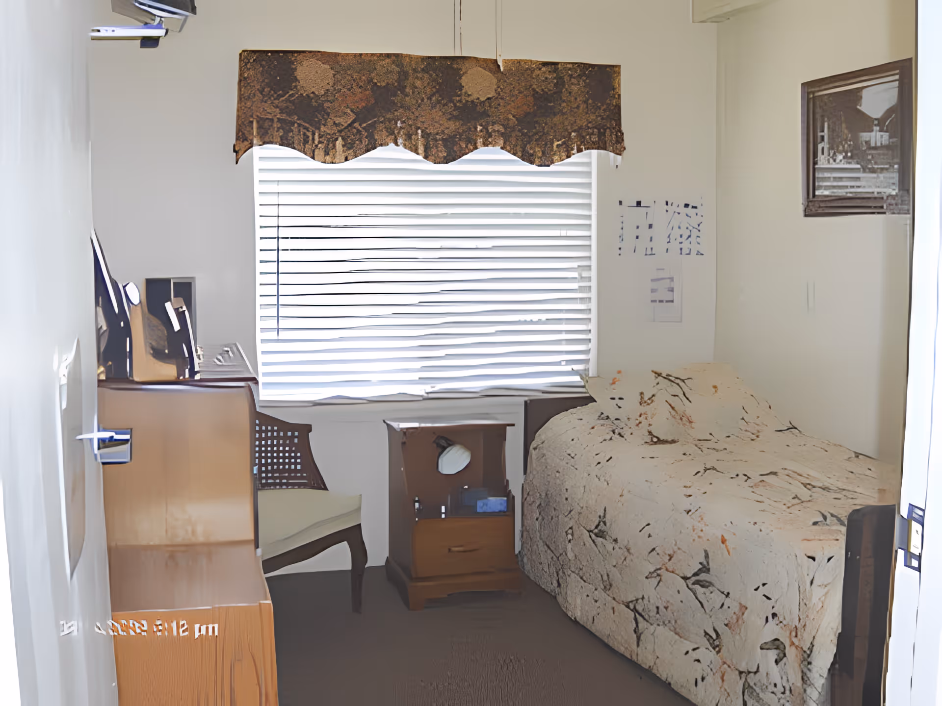 A small bedroom with a single bed covered in a floral quilt, a nightstand, desk, chair and a window with blinds.