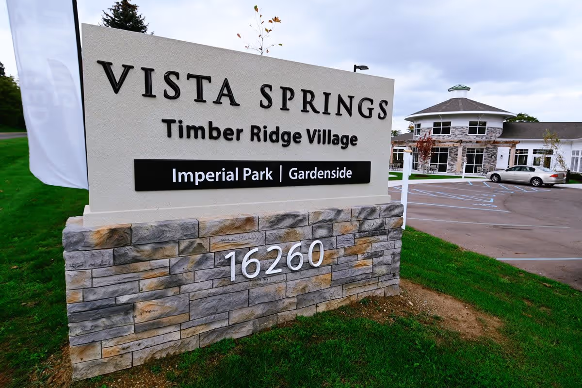 Outdoor view of the entrance sign for Vista Springs Timber Ridge Village, showing the address number 16260 and the names Imperial Park and Gardenside. In the background, there is a building with stone and white exterior and a parking lot with a car.