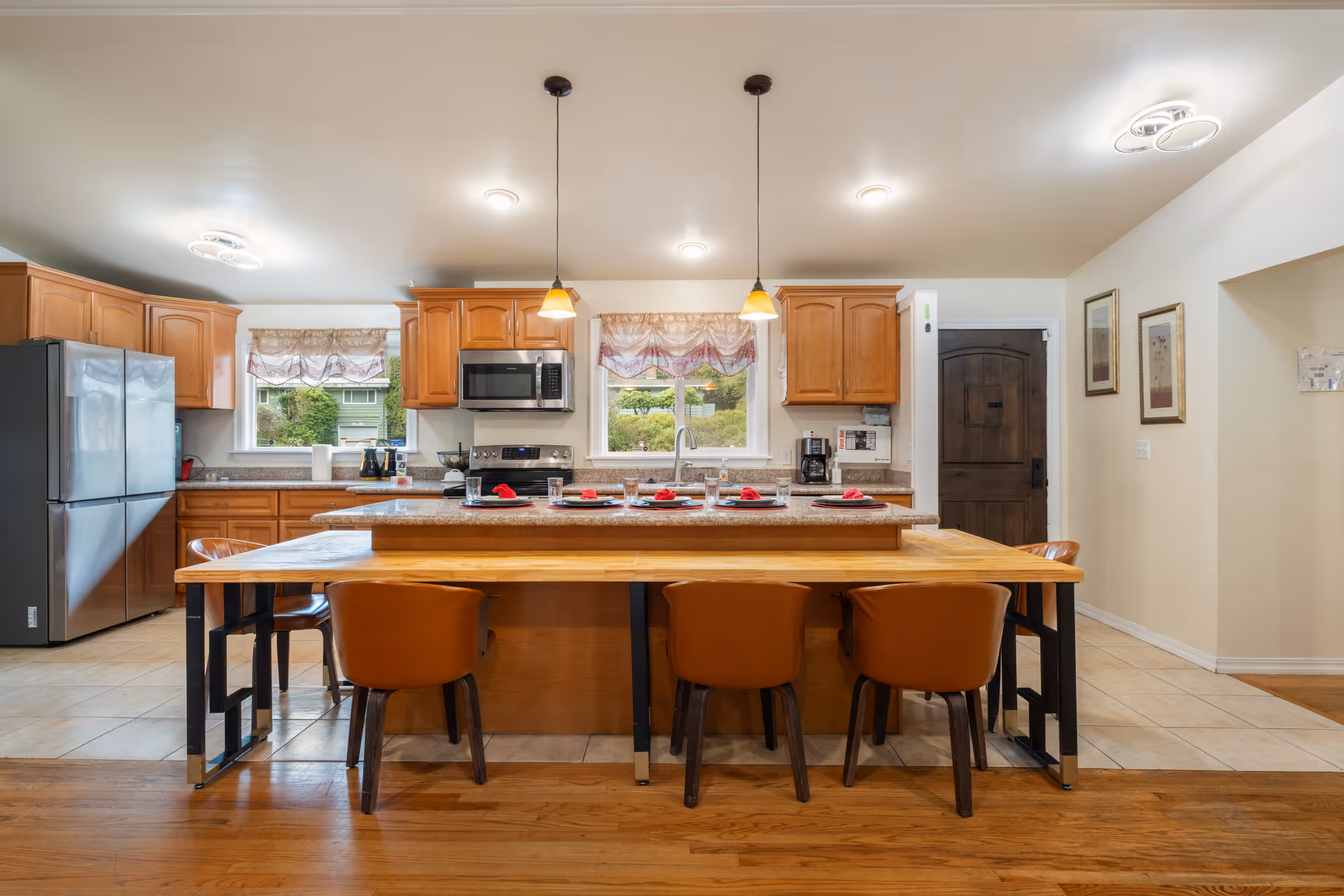 A spacious kitchen with wooden cabinets, a large island with a granite countertop, and a wooden dining table attached to the island. Four brown chairs are placed around the table. The kitchen features stainless steel appliances including a refrigerator, microwave, and stove. Two pendant lights hang above the island, and two windows with decorative curtains provide natural light. The floor is a combination of wood and tile, and there are framed pictures on the wall near a dark wooden door.