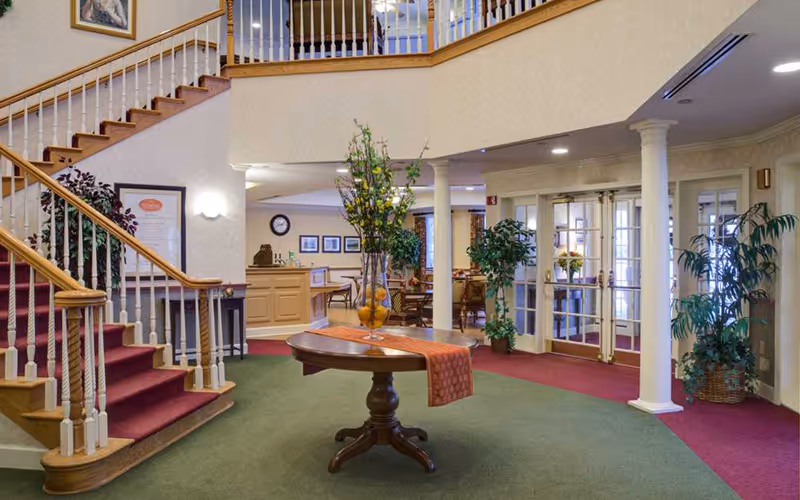 Bright senior living lobby with a central round table and floral arrangement, a staircase on the left, and seating and entrance doors in the background.