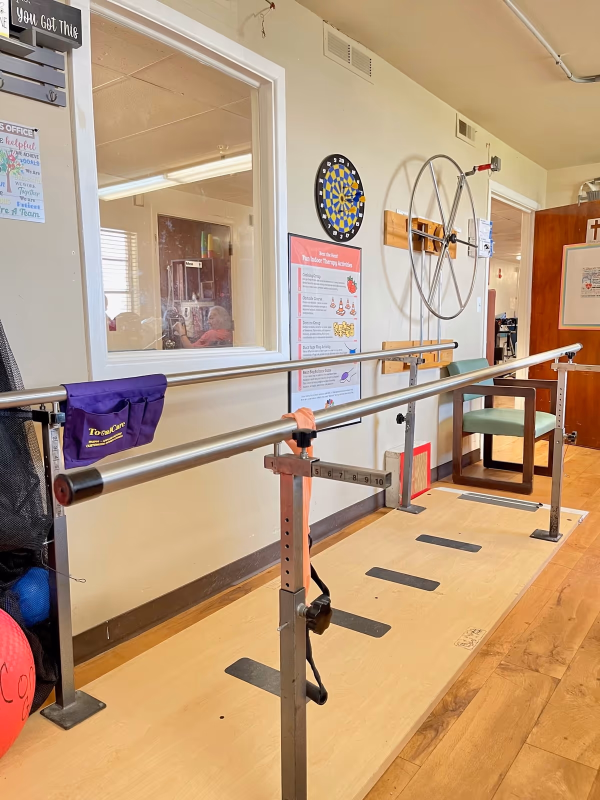 Physical therapy room with parallel bars for walking exercises, a dartboard on the wall, a large wheel mounted on the wall, a chair, and posters with therapy activities. The room has wooden flooring and light-colored walls.