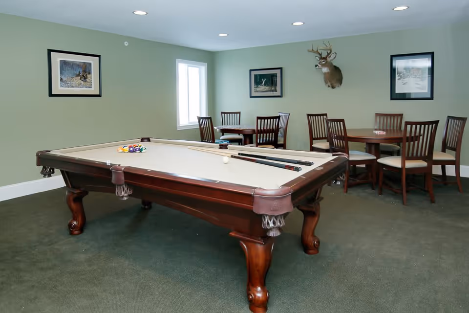 A recreational room with a pool table in the foreground, pool cues resting on the table, and billiard balls arranged for a game. In the background, there are two round wooden tables each surrounded by four chairs. The walls are painted light green and decorated with framed pictures and a mounted deer head. The room has a window letting in natural light and recessed ceiling lights.