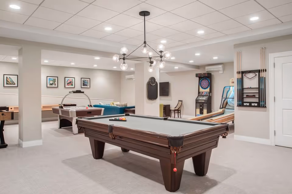 A spacious game room with a pool table in the center, an air hockey table, a foosball table, a dartboard, and a shuffleboard game. The room has light-colored walls and carpet, recessed lighting, and a modern chandelier above the pool table. There is a seating area with a blue couch and framed artwork on the walls.