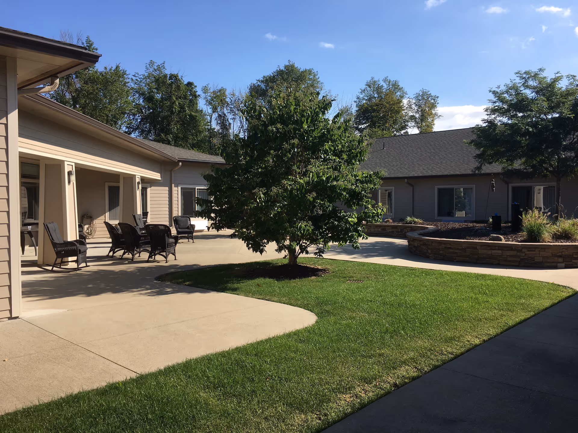 Outdoor courtyard area at Windsor Heights Assisted Living and Memory Care with a central tree, green grass, paved walkways, and patio seating with black chairs under a covered porch. The building surrounds the courtyard with windows and doors visible.