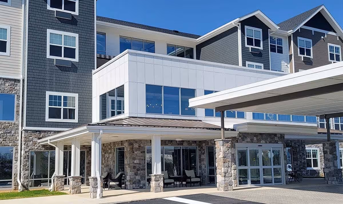 Exterior view of a modern senior living facility building with stone and siding facade, large windows, and a covered entrance with seating area under a clear blue sky.