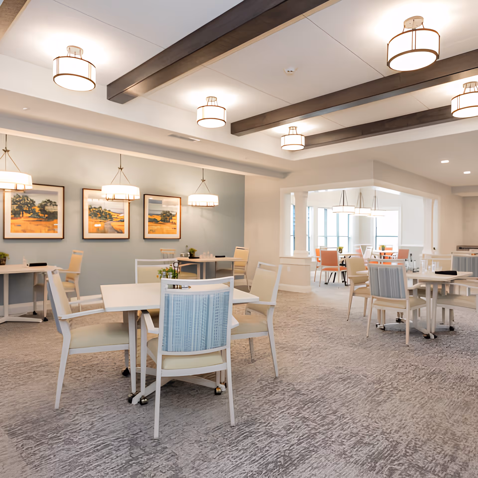 A bright and spacious dining area in a senior living facility with multiple tables and chairs arranged neatly. The room features modern light fixtures hanging from the ceiling with wooden beams, framed landscape artwork on the walls, and a carpeted floor. There is an adjoining room visible in the background with additional seating and large windows allowing natural light to enter.
