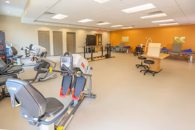 A spacious rehabilitation or physical therapy room with exercise equipment including stationary bikes and leg press machines, parallel bars for walking practice, therapy tables, a wheelchair, and stools. The room has a bright yellow accent wall and several closed doors.