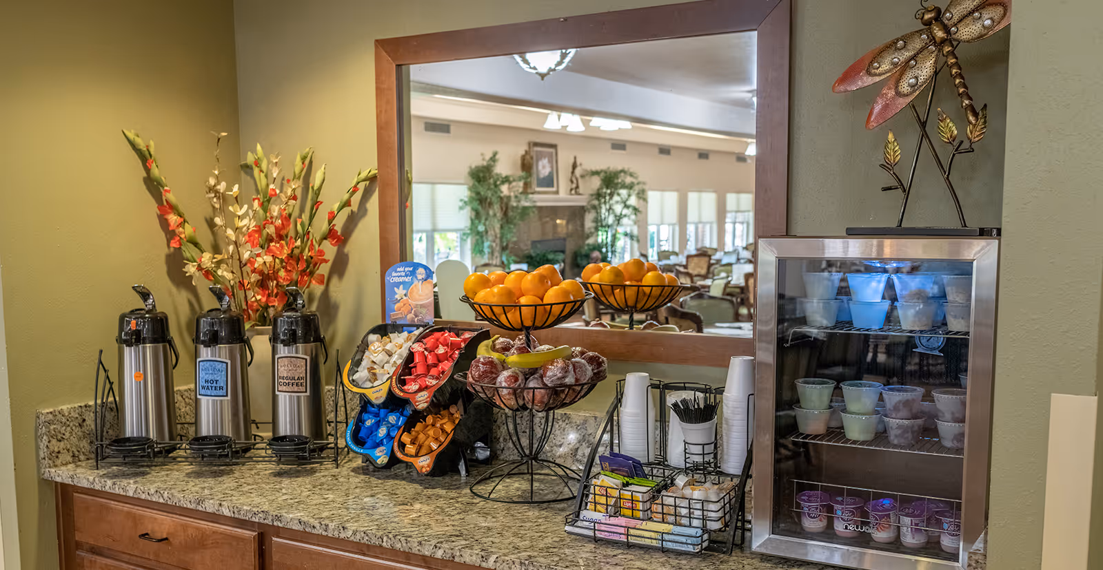 A coffee and snack station with three stainless steel dispensers labeled hot water and regular coffee, a variety of coffee creamers and sweeteners, two wire baskets filled with oranges and wrapped pastries, disposable cups and stirrers, and a small refrigerated display case with assorted yogurt cups. A large mirror behind the station reflects a spacious lounge area with chairs, tables, plants, and a fireplace.