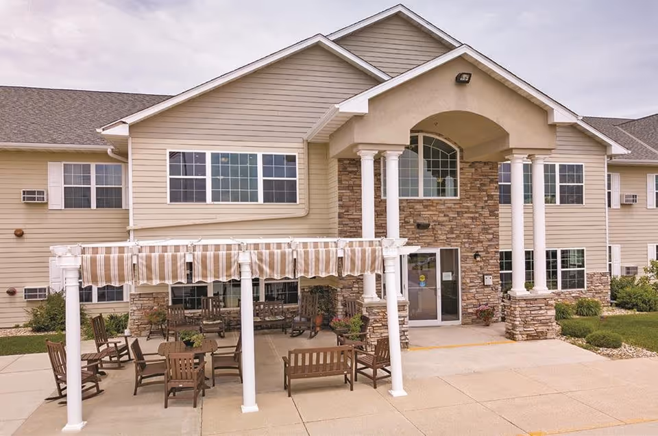 Exterior view of a senior living facility named Keelson Harbour featuring a beige building with stone accents, large windows, and a covered entrance supported by white columns. In front of the building, there is a patio area with wooden chairs and tables under a striped canopy.