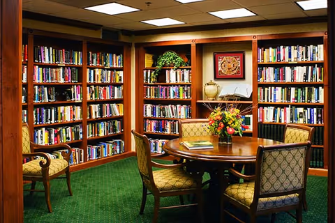 A cozy library room with wooden bookshelves filled with books lining the walls. In the center, there is a round wooden table with a floral arrangement and several chairs with patterned upholstery around it. The room has green carpet and warm lighting from ceiling fixtures.