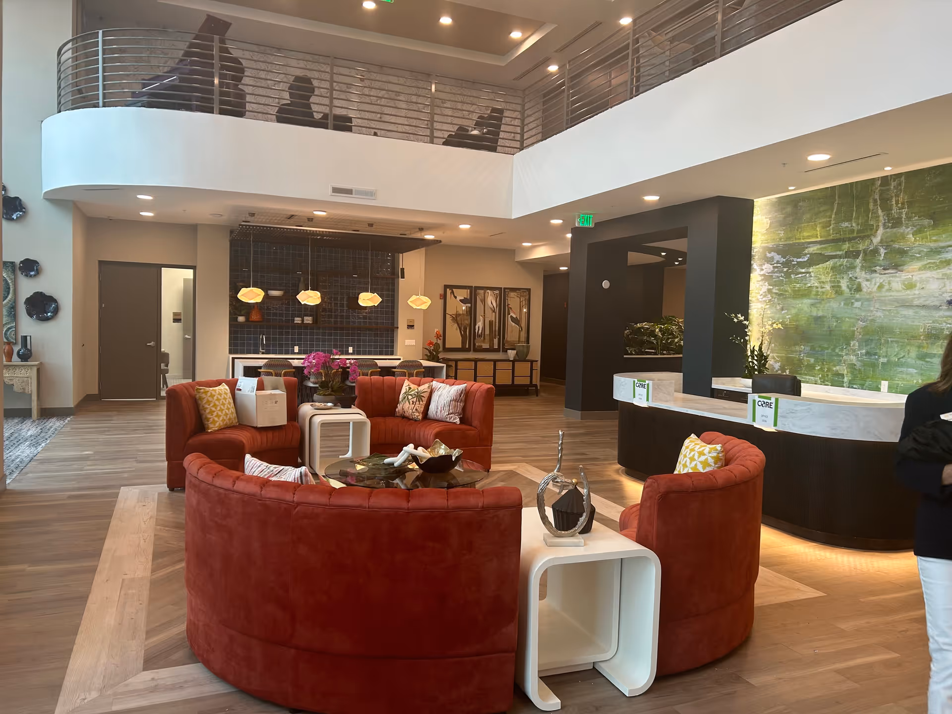 A modern senior living facility lobby with a circular red sofa set around a glass coffee table, decorative pillows, and a white side table. The space features wood flooring, a reception desk with a green textured wall behind it, and a mezzanine level with metal railings. Pendant lights hang over a counter area in the background, and there are decorative wall pieces and plants throughout.